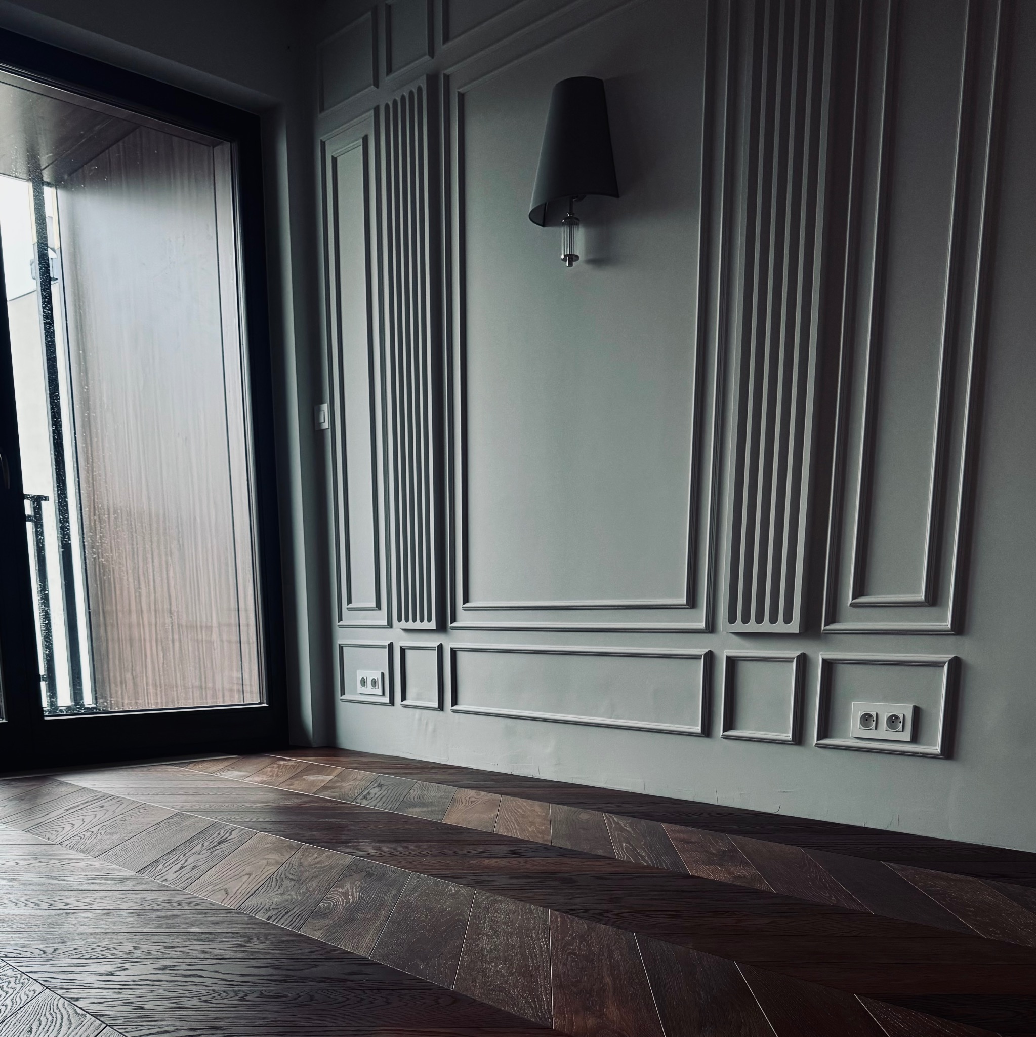 Ułożona w jodełkę, ciemna, drewniana podłoga w nowocześnie urządzonym wnętrzu z szarą ścianą, dekoracyjnymi listwami i czarnym oknem balkonowym.