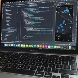 Laptop MacBook Pro z otwartym edytorem kodu (Flutter/Dart) i uruchomionym emulatorem Androida z mapą i pinezkami lokalizacji, prawdopodobnie w trakcie tworzenia aplikacji mobilnej.