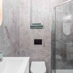 Enjoy Construction Paweł Kornecki - Nowoczesna łazienka z szarymi płytkami imitującymi kamień, kabiną prysznicową ze szklanymi drzwiami, białym grzejnikiem drabinkowym, umywalką z chromowaną baterią i okrągłym lustrem z drewnianą ramą.