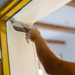 Nakładanie gładzi szpachlowej na ścianę przy żółtej futrynie drzwi, widoczna dłoń z pacą, użyta niebieska taśma malarska