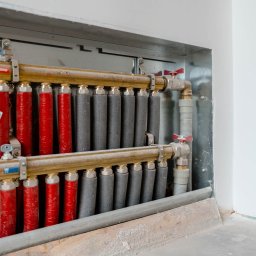 Enjoy Construction Paweł Kornecki - Rozdzielacz ogrzewania podłogowego z rurami w izolacji czerwonej i szarej, zamontowany w metalowej obudowie w trakcie prac wykończeniowych.