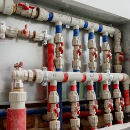 Enjoy Construction Paweł Kornecki - Rozdzielacz wodny z rurami PEX w kolorach czerwonym i niebieskim, zamontowany na metalowej płycie w betonowej wnęce, widoczne zawory z czerwonymi uchwytami.
