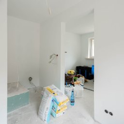 Enjoy Construction Paweł Kornecki - Wnętrze w trakcie prac wykończeniowych: świeżo pomalowane białe ściany, worki z zaprawą gipsową Stais SMS 15, narzędzia malarskie i wystające przewody elektryczne.