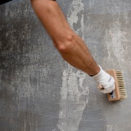 Enjoy Construction Paweł Kornecki - Szorstka, nierówna ściana w odcieniach szarości, czyszczona szczotką przez osobę w białej rękawicy, widoczne ślady wcześniejszych warstw i nierówności.