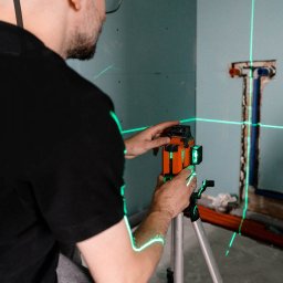 Enjoy Construction Paweł Kornecki - Instalator ustawia laser krzyżowy na statywie, wyznaczając pion i poziom na ścianie z przygotowanymi przyłączami hydraulicznymi.