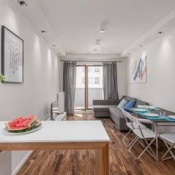 Enjoy Construction Paweł Kornecki - Jasne wnętrze mieszkania z drewnianą podłogą, szarą sofą, stołem z nakryciem i wazonem z kwiatami oraz talerzem arbuza na blacie