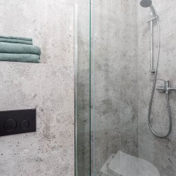 Enjoy Construction Paweł Kornecki - Nowoczesna łazienka z kabiną prysznicową, szarymi płytkami imitującymi kamień, czarnym przyciskiem spłuczki i stosem turkusowych ręczników wnęce.