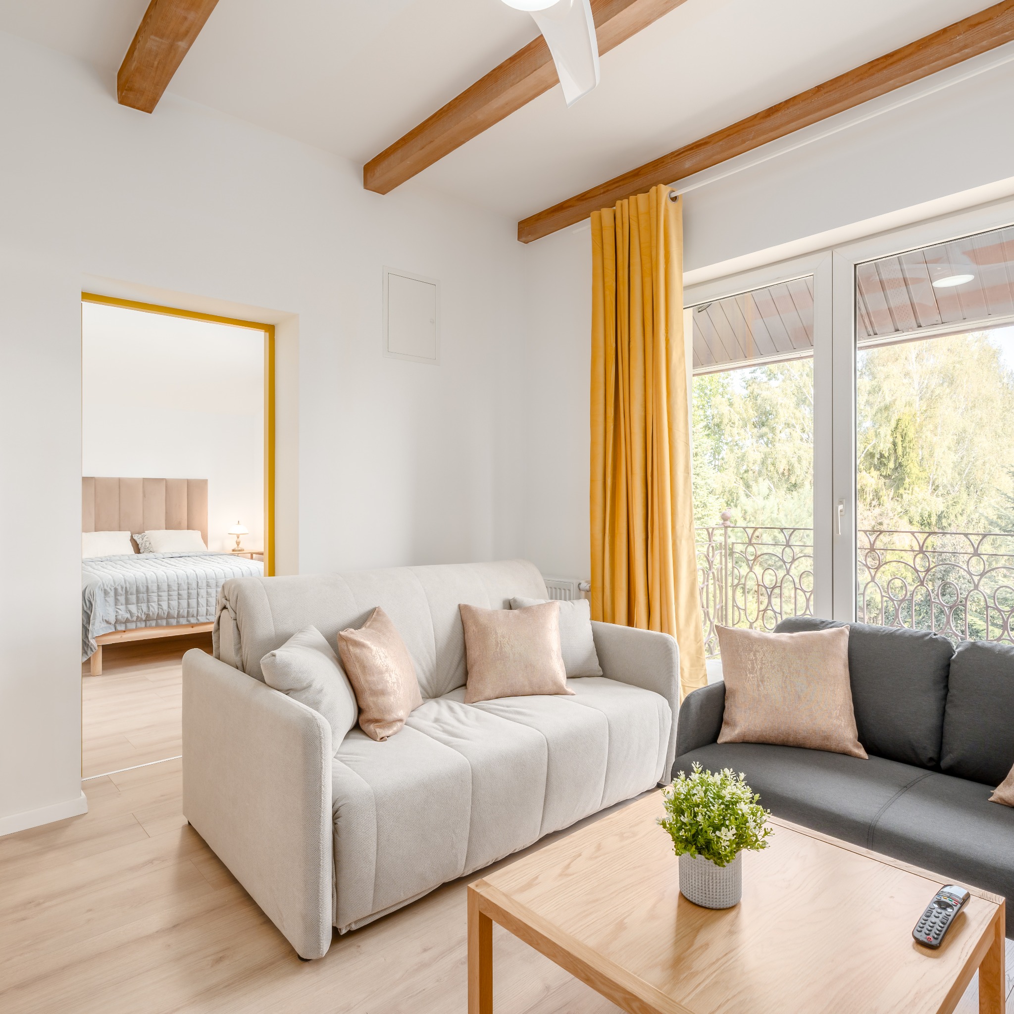 Jasne wnętrze pokoju dziennego z beżową sofą, drewnianymi belkami stropowymi, żółtymi zasłonami i widokiem na sypialnię przez otwarte drzwi.