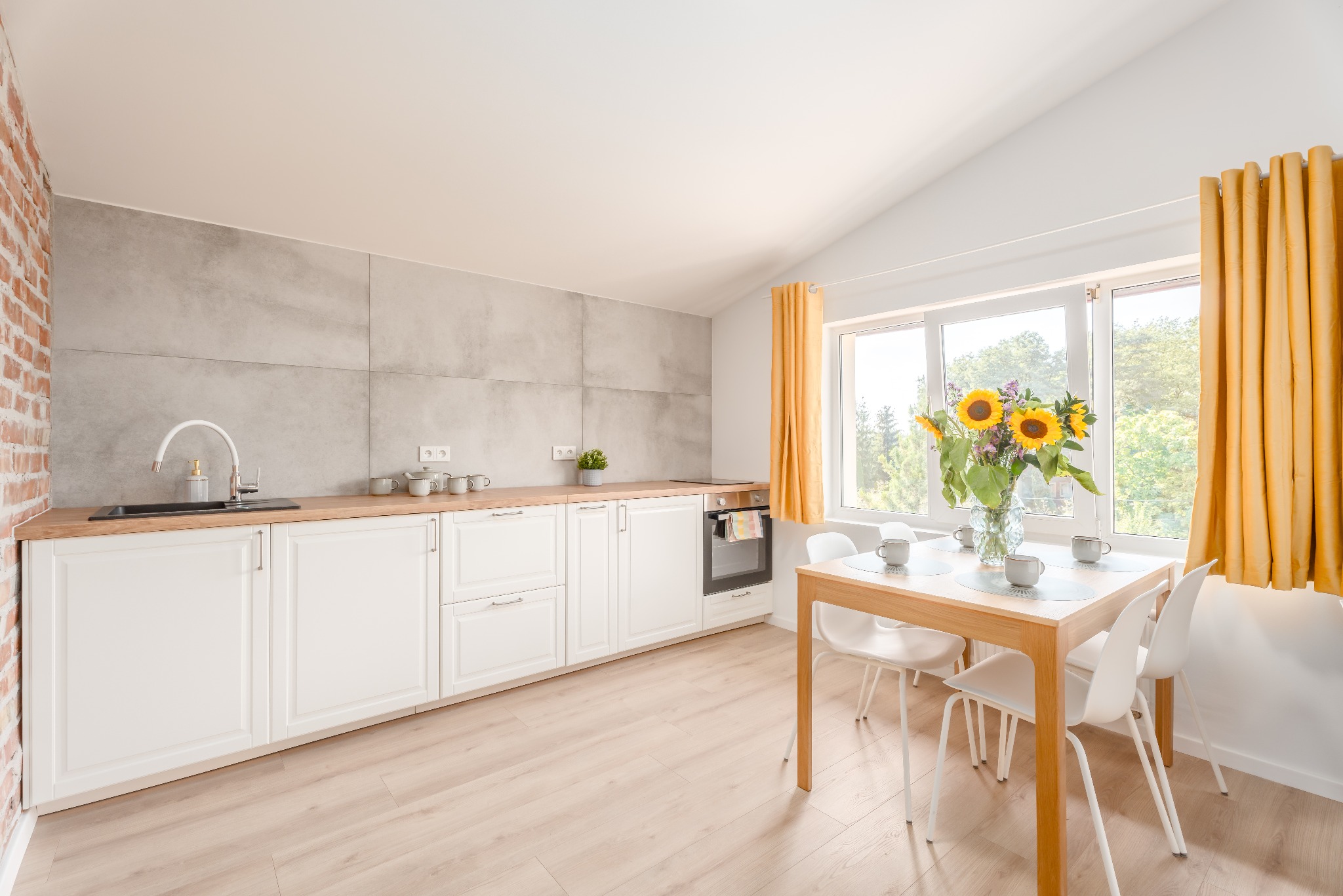 Jasna kuchnia z ceglaną ścianą, szarymi panelami, białymi szafkami, drewnianym blatem i stołem z bukietem słoneczników.