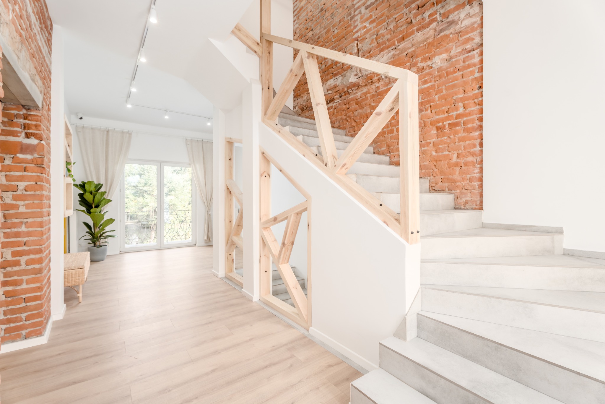 Nowoczesne schody z drewnianą balustradą i szarymi stopniami, ceglana ściana w tle, parkiet na podłodze, jasne wnętrze z rośliną w doniczce.