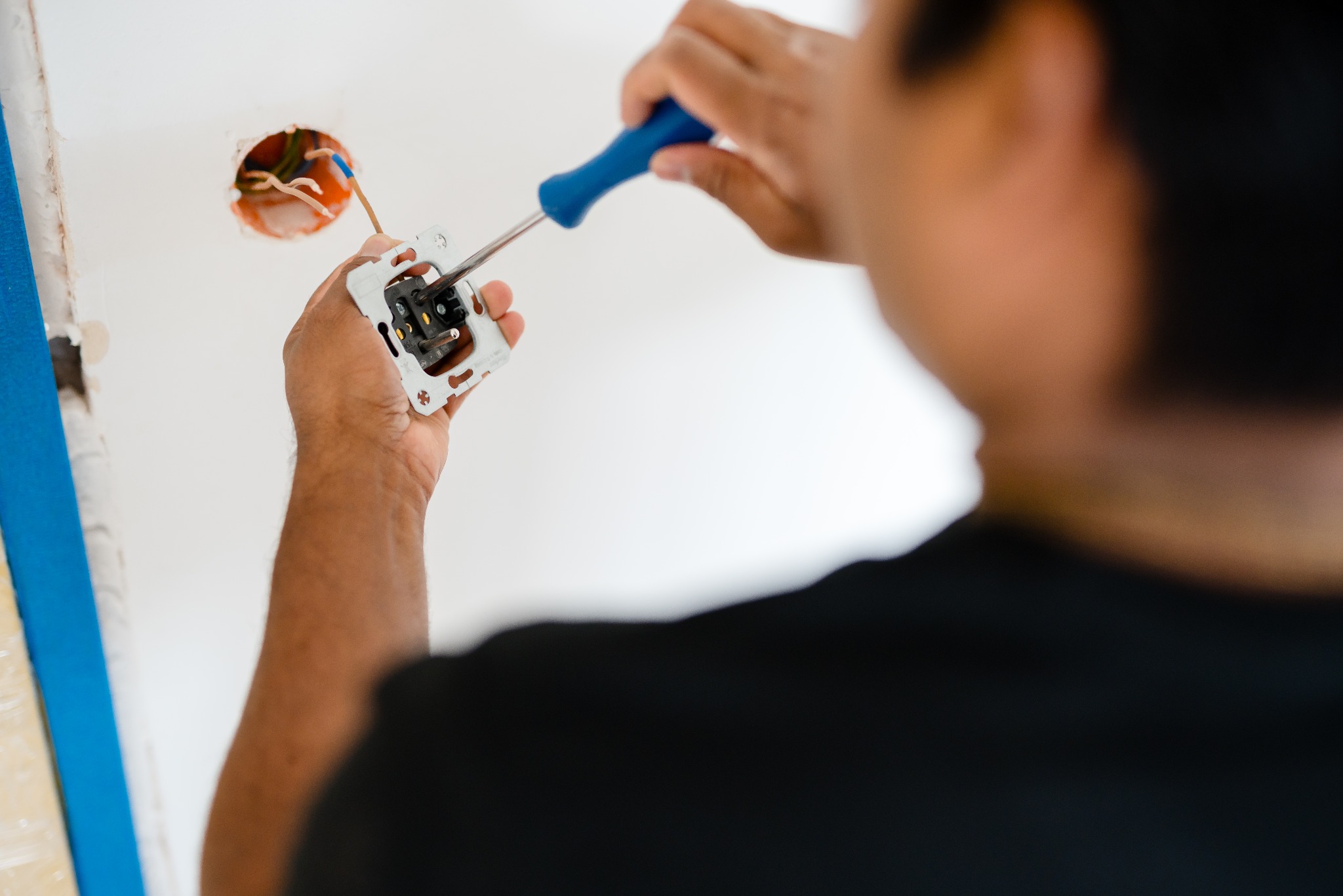 Elektryk z niebieskim śrubokrętem montuje gniazdko elektryczne w ścianie, widoczna skrzynka instalacyjna z przewodami i taśma malarska zabezpieczająca ścianę.