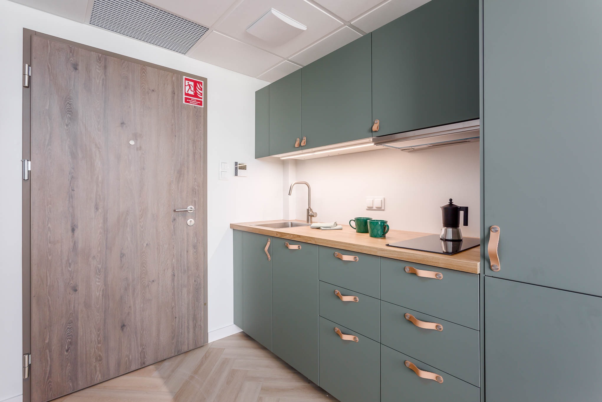 Nowoczesna, zielona kuchnia z drewnianym blatem i skórzanymi uchwytami, widok na drzwi z oznaczeniem przeciwpożarowym i parkiet ułożony w jodełkę.