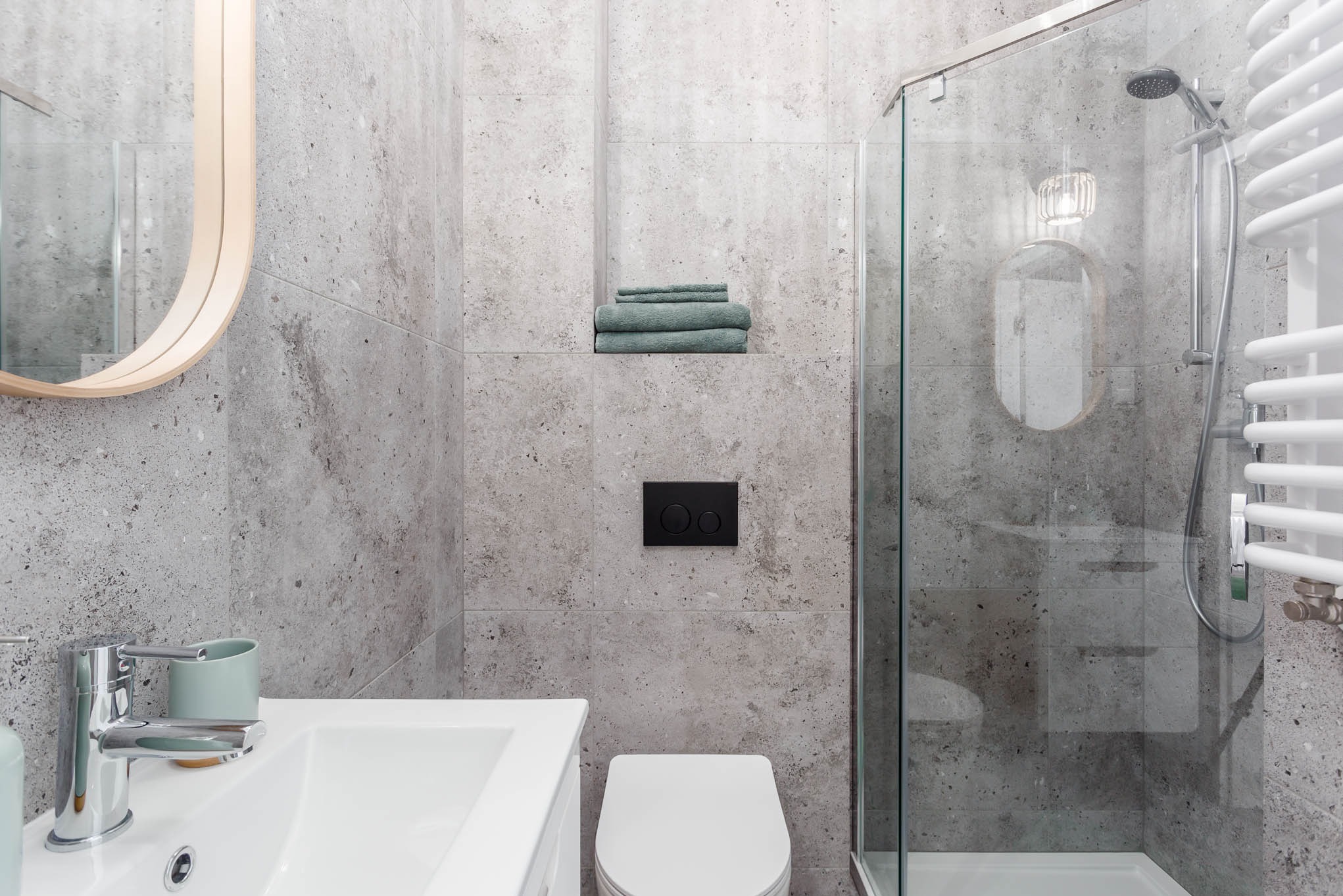 Nowoczesna łazienka z szarymi płytkami imitującymi kamień, kabiną prysznicową ze szklanymi drzwiami, białym grzejnikiem drabinkowym, umywalką z chromowaną baterią i okrągłym lustrem z drewnianą ramą.