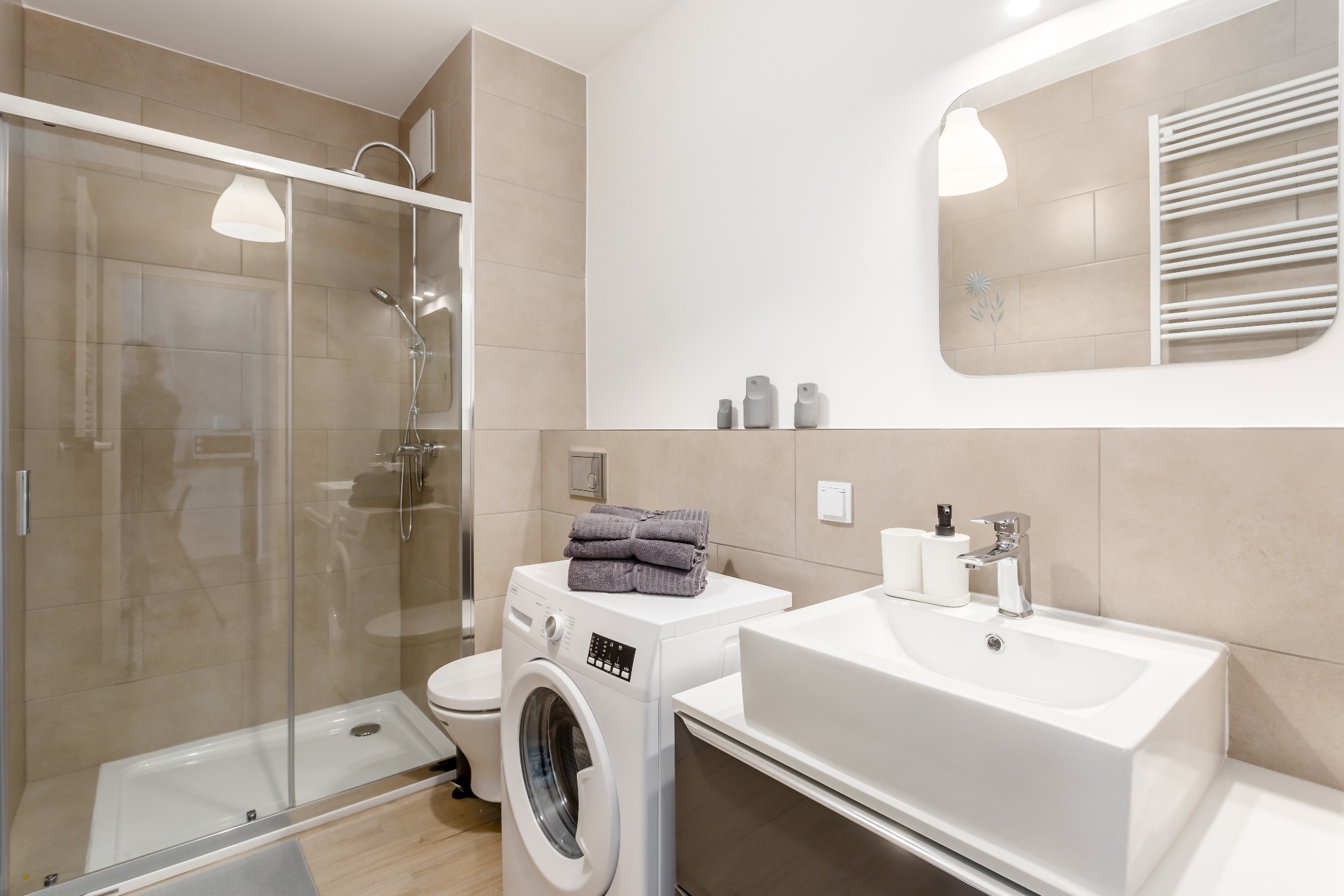 Jasna łazienka z kabiną prysznicową, pralką z ułożonymi ręcznikami i umywalką nablatową, wykończona beżowymi płytkami.