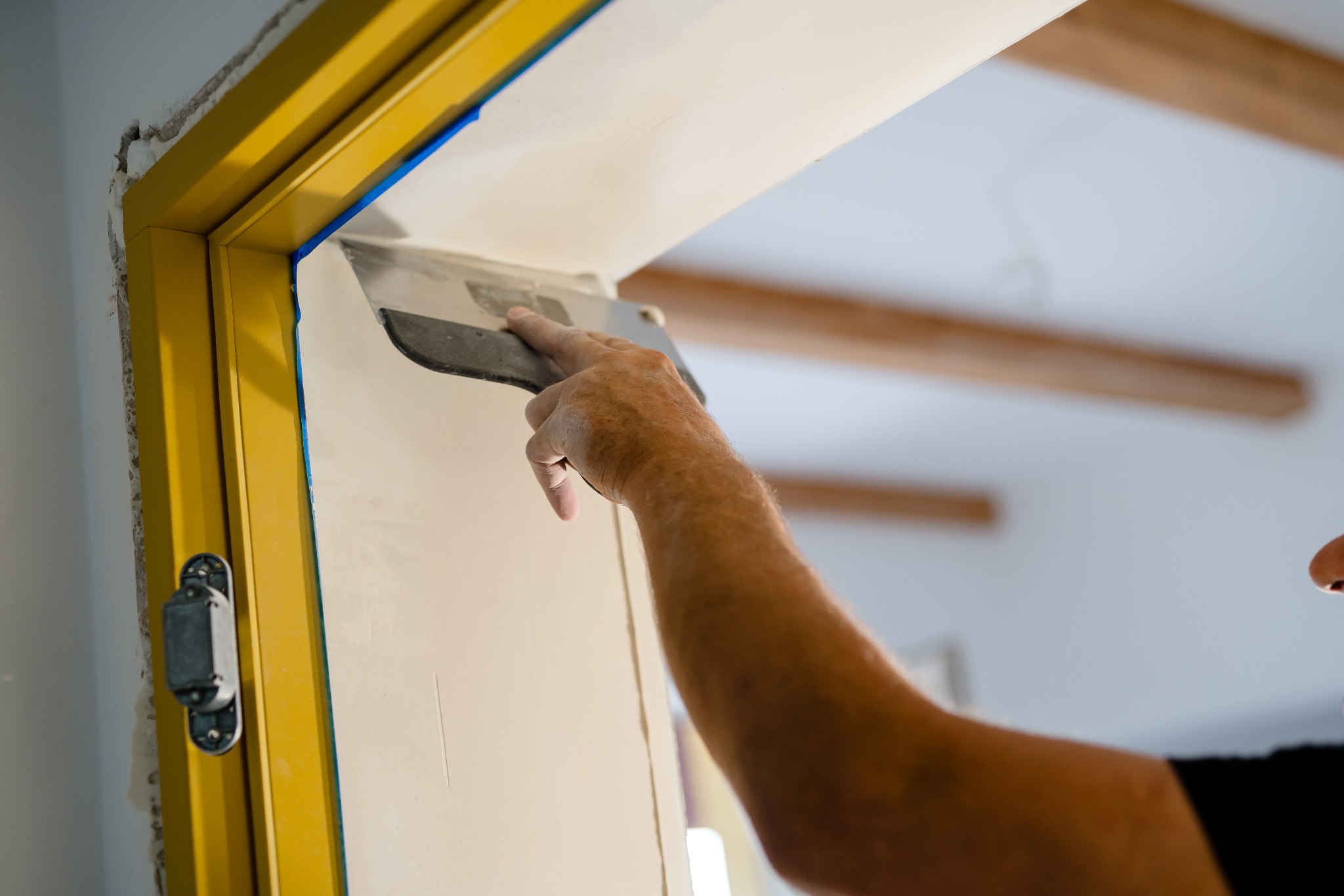 Nakładanie gładzi szpachlowej na ścianę przy żółtej futrynie drzwi, widoczna dłoń z pacą, użyta niebieska taśma malarska