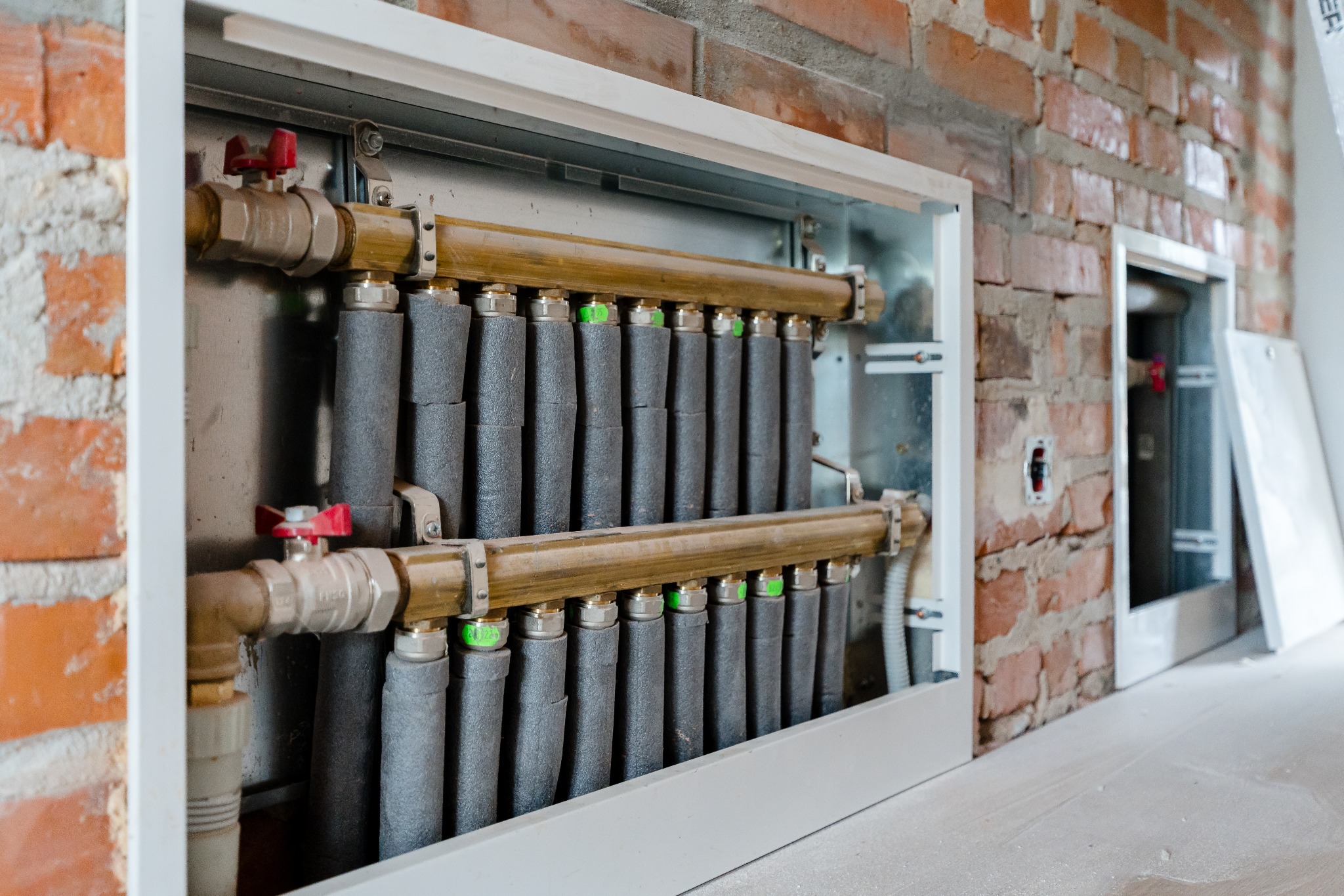 Rozdzielacz ogrzewania podłogowego z izolowanymi rurami, zamontowany w szafce w surowej ścianie z czerwonej cegły, widoczne fragmenty instalacji elektrycznej i otwarta szafka obok.