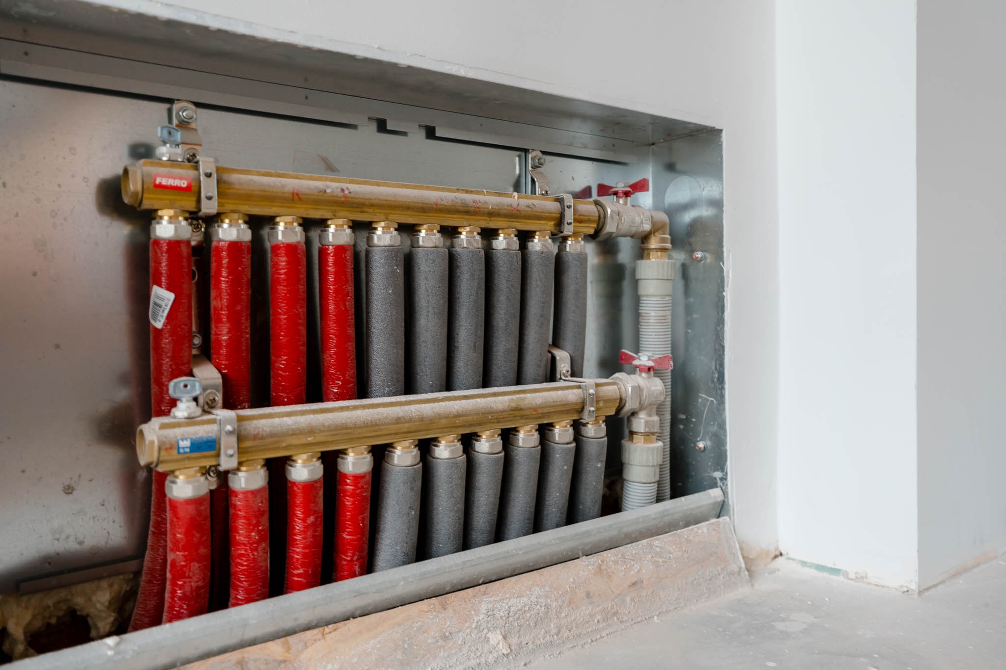 Rozdzielacz ogrzewania podłogowego z rurami w izolacji czerwonej i szarej, zamontowany w metalowej obudowie w trakcie prac wykończeniowych.