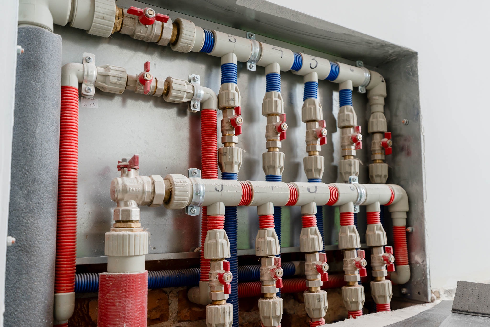 Rozdzielacz wodny z rurami PEX w kolorach czerwonym i niebieskim, zamontowany na metalowej płycie w betonowej wnęce, widoczne zawory z czerwonymi uchwytami.