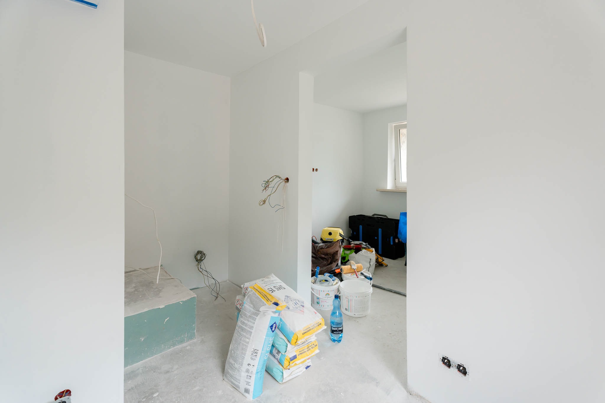 Wnętrze w trakcie prac wykończeniowych: świeżo pomalowane białe ściany, worki z zaprawą gipsową Stais SMS 15, narzędzia malarskie i wystające przewody elektryczne.