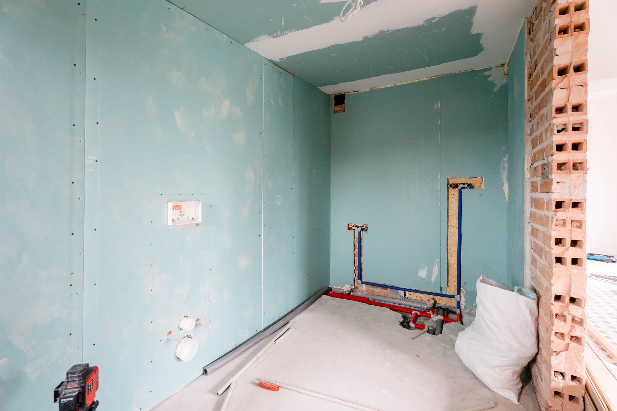 Wnętrze łazienki w trakcie prac wykończeniowych: ściany pokryte płytami kartonowo-gipsowymi w odcieniach błękitu, widoczne instalacje wodne i narzędzia na podłodze, fragment ściany z cegły dziurawki.