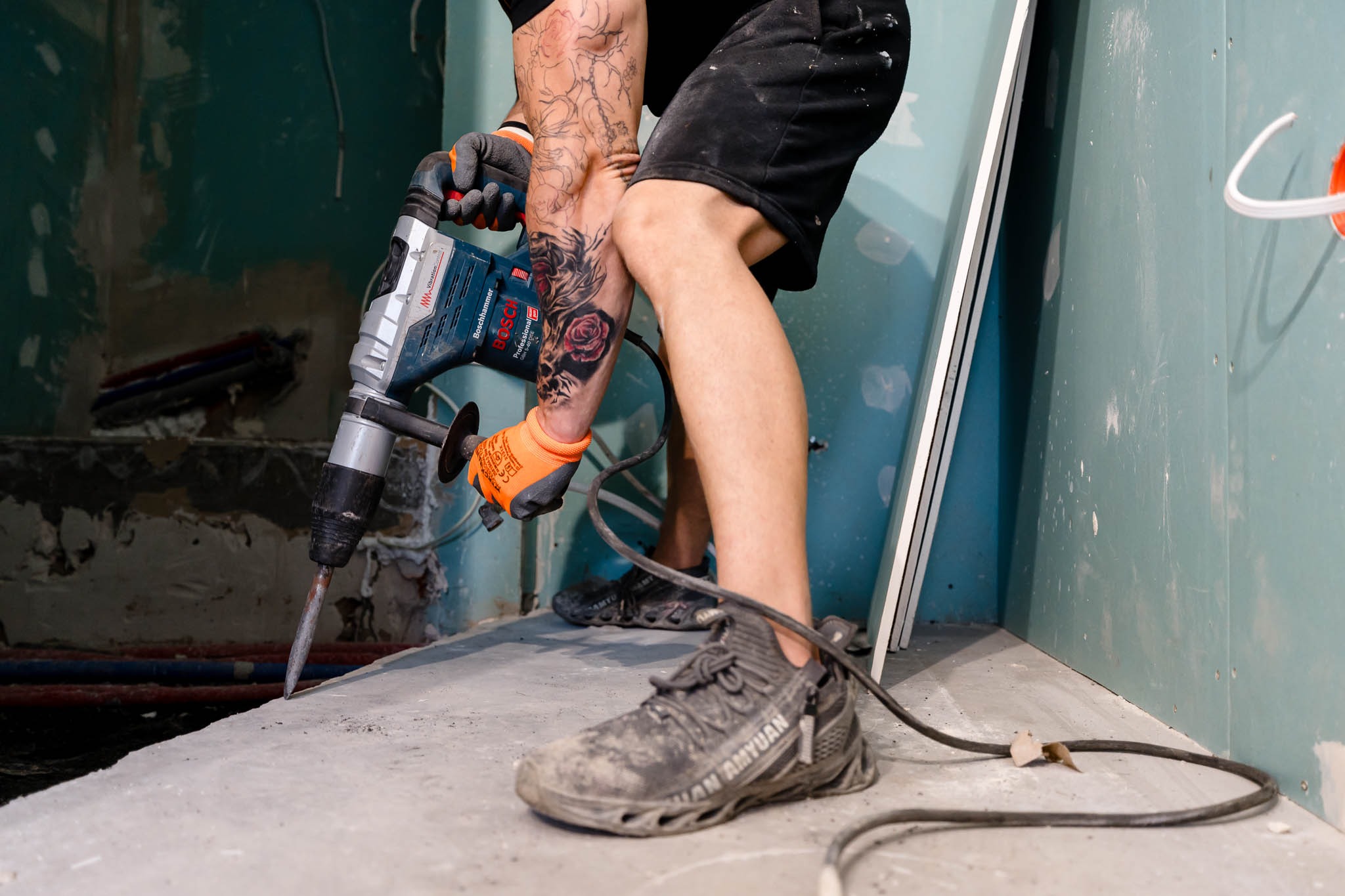 Pracownik z tatuażami, w rękawicach i roboczych butach, używa młota udarowego Bosch na podłodze w pomieszczeniu z częściowo wykończonymi ścianami z płyty karton gips.