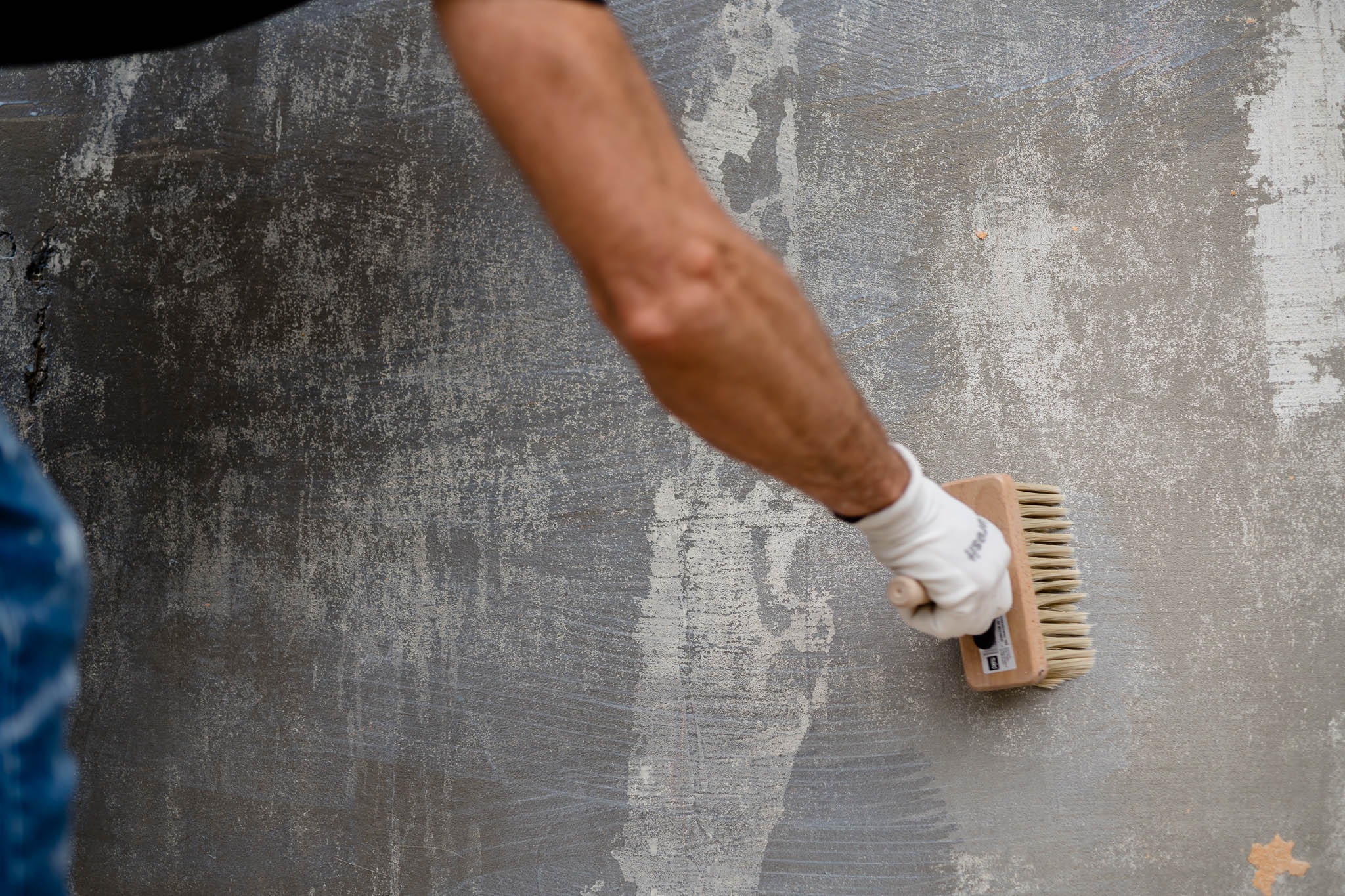 Szorstka, nierówna ściana w odcieniach szarości, czyszczona szczotką przez osobę w białej rękawicy, widoczne ślady wcześniejszych warstw i nierówności.