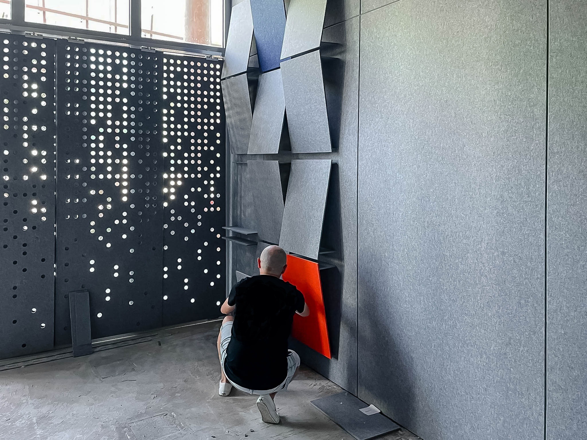 Montaż paneli akustycznych na ścianie z dekoracyjnym ażurem, widoczny pracownik w trakcie mocowania pomarańczowego panelu.