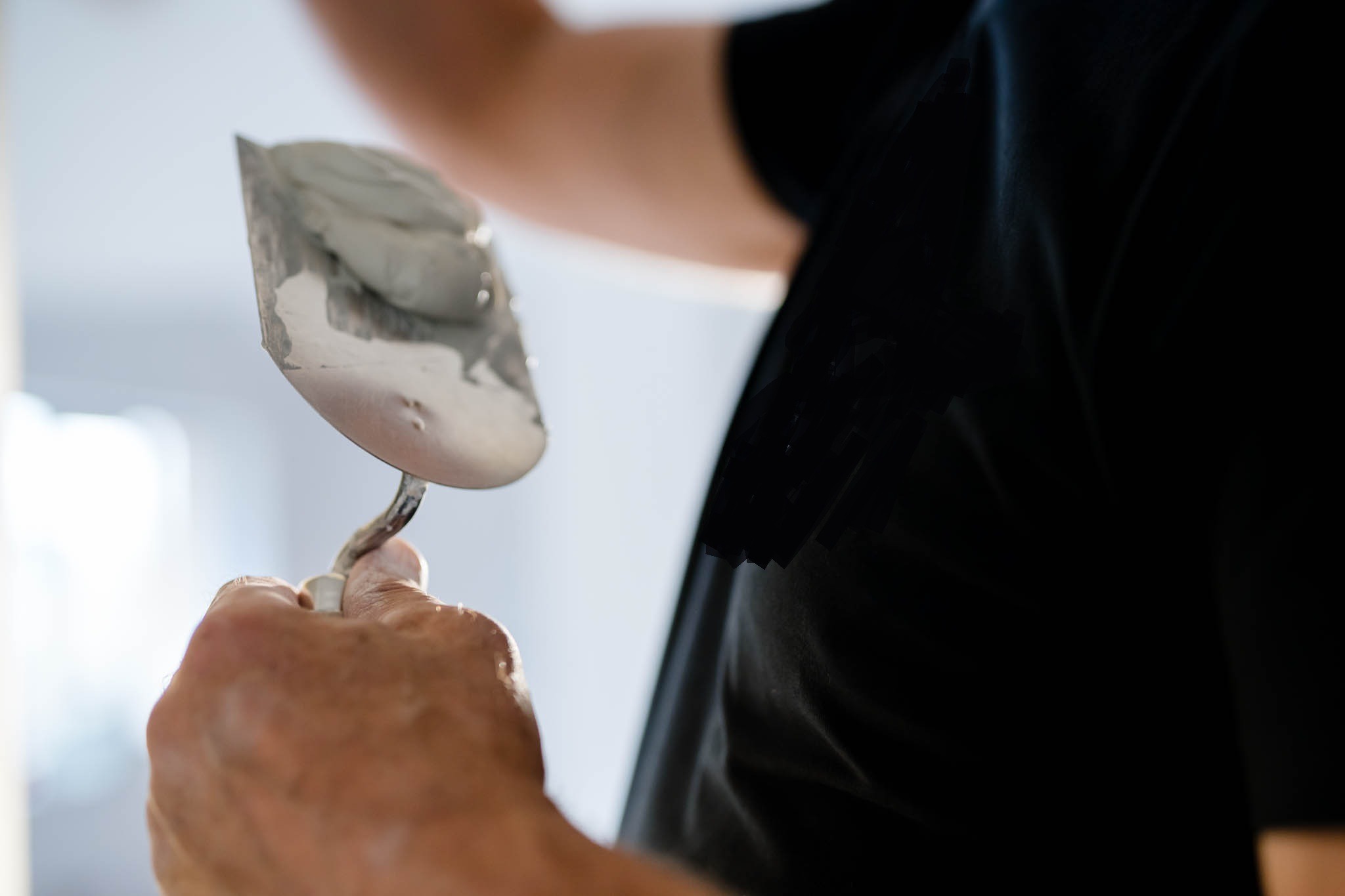 Szpachelka z nałożoną szarą masą w dłoni pracownika ubranego w czarną koszulkę, przygotowanie do rozprowadzania masy.