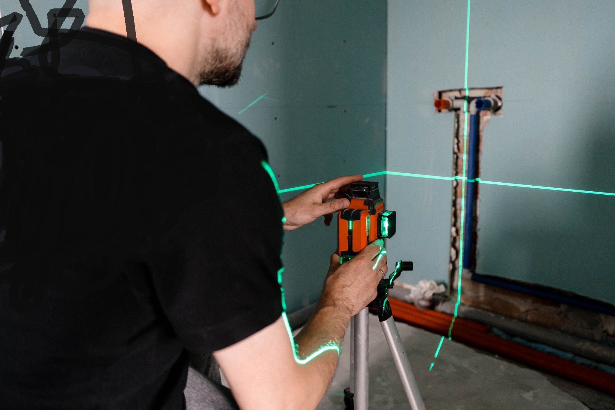 Instalator ustawia laser krzyżowy na statywie, wyznaczając pion i poziom na ścianie z przygotowanymi przyłączami hydraulicznymi.