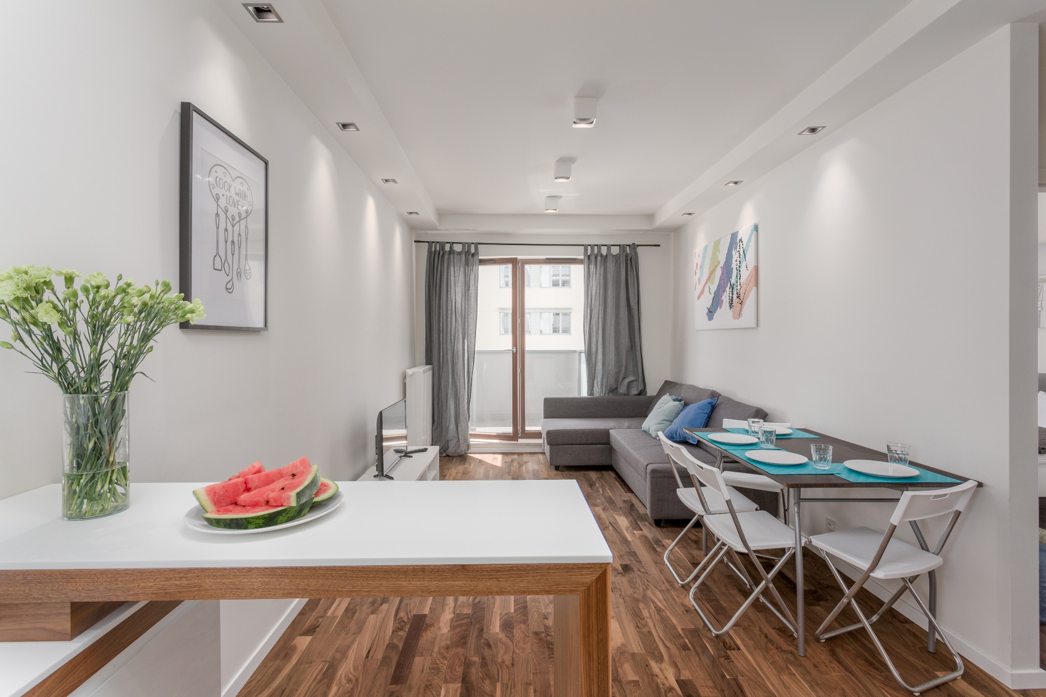 Jasne wnętrze mieszkania z drewnianą podłogą, szarą sofą, stołem z nakryciem i wazonem z kwiatami oraz talerzem arbuza na blacie