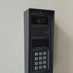 Szary domofon Elfon z klawiaturą numeryczną i czytnikiem kart zbliżeniowych, zamontowany na jasnej ścianie. Widoczne przyciski i instrukcja 'W razie pomyłki naciśnij #'.