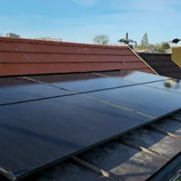 Cztery moduły fotowoltaiczne, zamontowane na dachu z czerwoną dachówką, w słoneczny dzień. Widoczne elementy montażowe i fragmenty konstrukcji dachu.