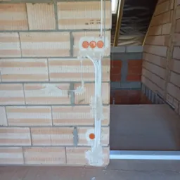 Wewnętrzna instalacja elektryczna w trakcie montażu: przewody w ścianie z cegły, pomarańczowe puszki pod gniazdka, surowy stan wykończenia wnętrza.