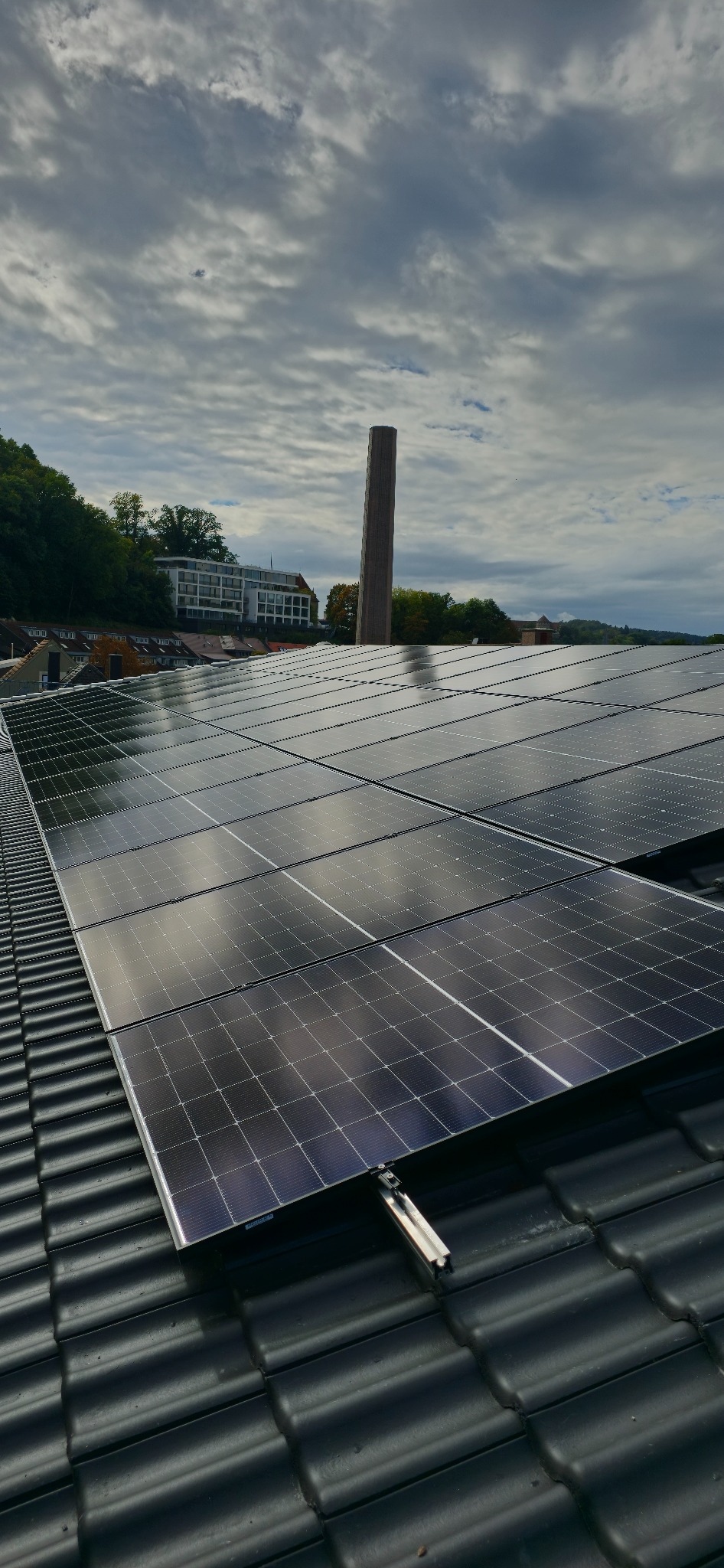 Rząd paneli fotowoltaicznych na czarnym dachu z dachówki ceramicznej, w tle budynek i komin na tle pochmurnego nieba.