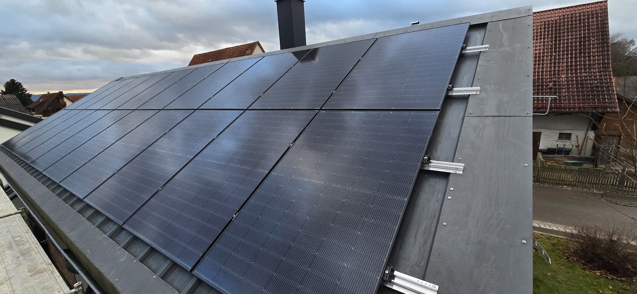 Czarny panel fotowoltaiczny zamontowany na szarym dachu, widoczne mocowania. W tle zabudowa jednorodzinna i pochmurne niebo.