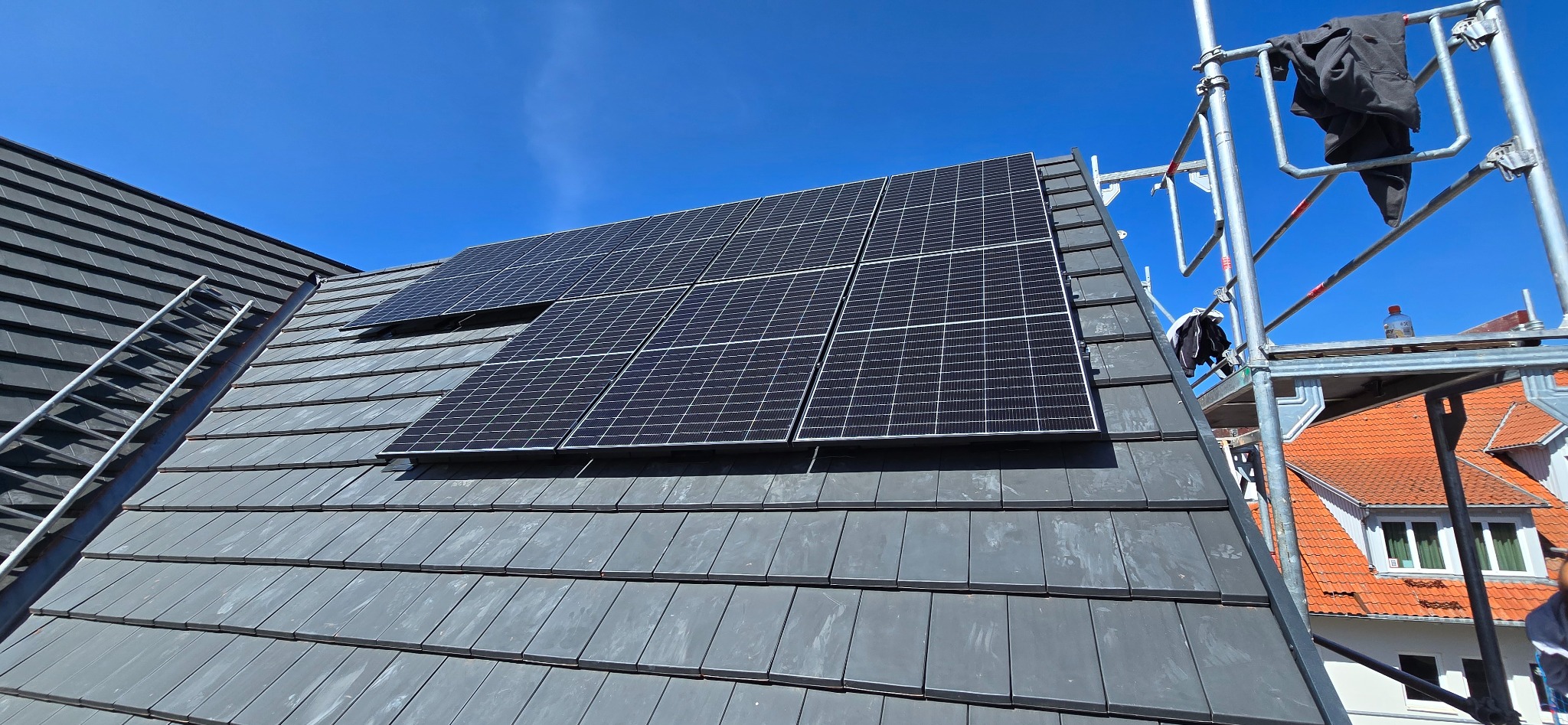 Panele fotowoltaiczne na dachu z szarej dachówki, widoczne rusztowanie i drabina. Błękitne niebo w tle, częściowo widoczny budynek z czerwoną dachówką.