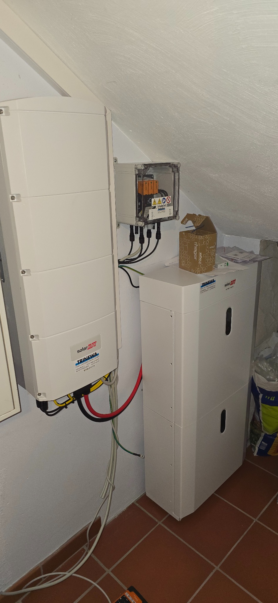 Biały falownik SolarEdge i magazyn energii Trinewa zamontowane na ścianie w pomieszczeniu ze skośnym sufitem. Widoczne kable i skrzynka zabezpieczeń.