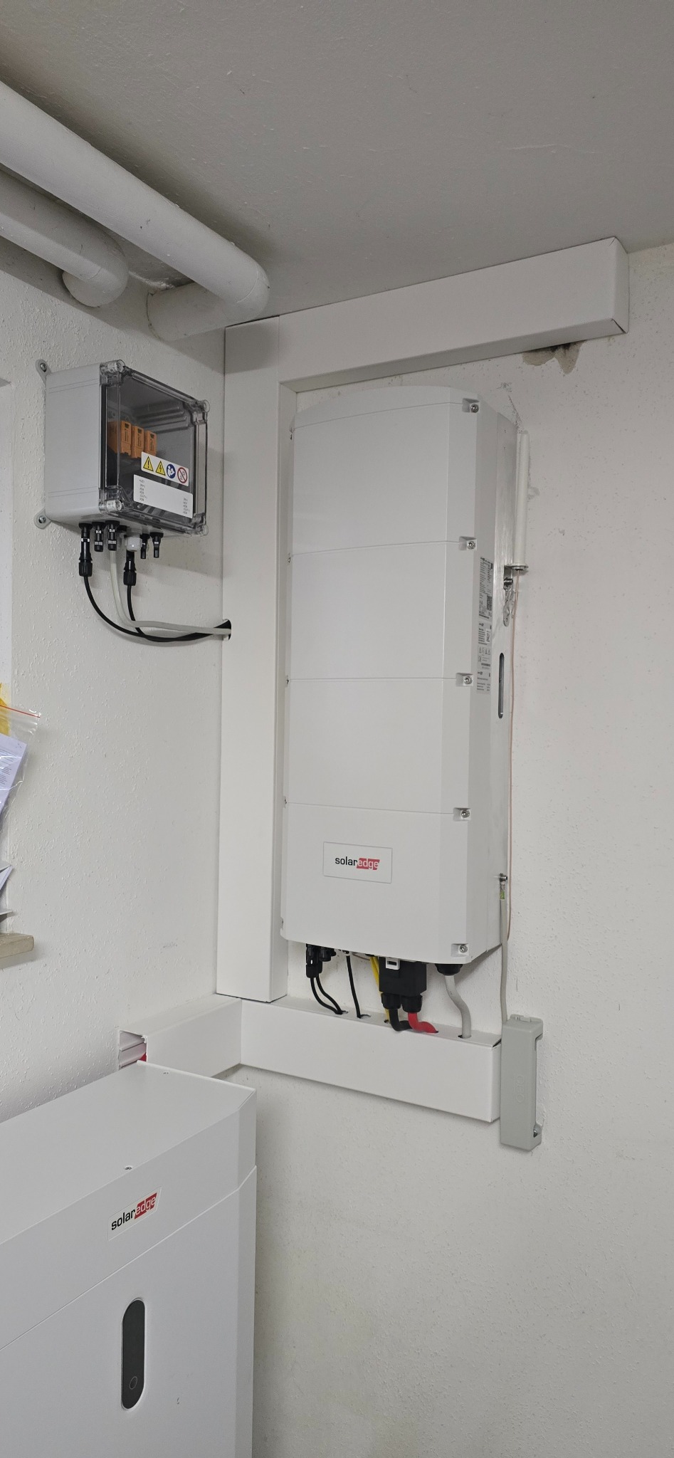 Biały falownik SolarEdge zamontowany na ścianie wewnątrz budynku, z widoczną skrzynką bezpiecznikową i przewodami. Element instalacji fotowoltaicznej.