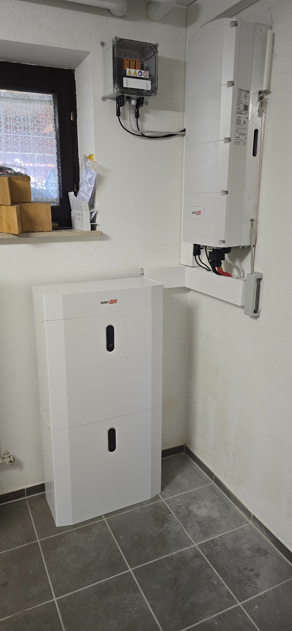 Biały, dwuczęściowy magazyn energii SolarEdge zamontowany na ścianie w pomieszczeniu z szarą podłogą. Widoczna skrzynka elektryczna i okno.