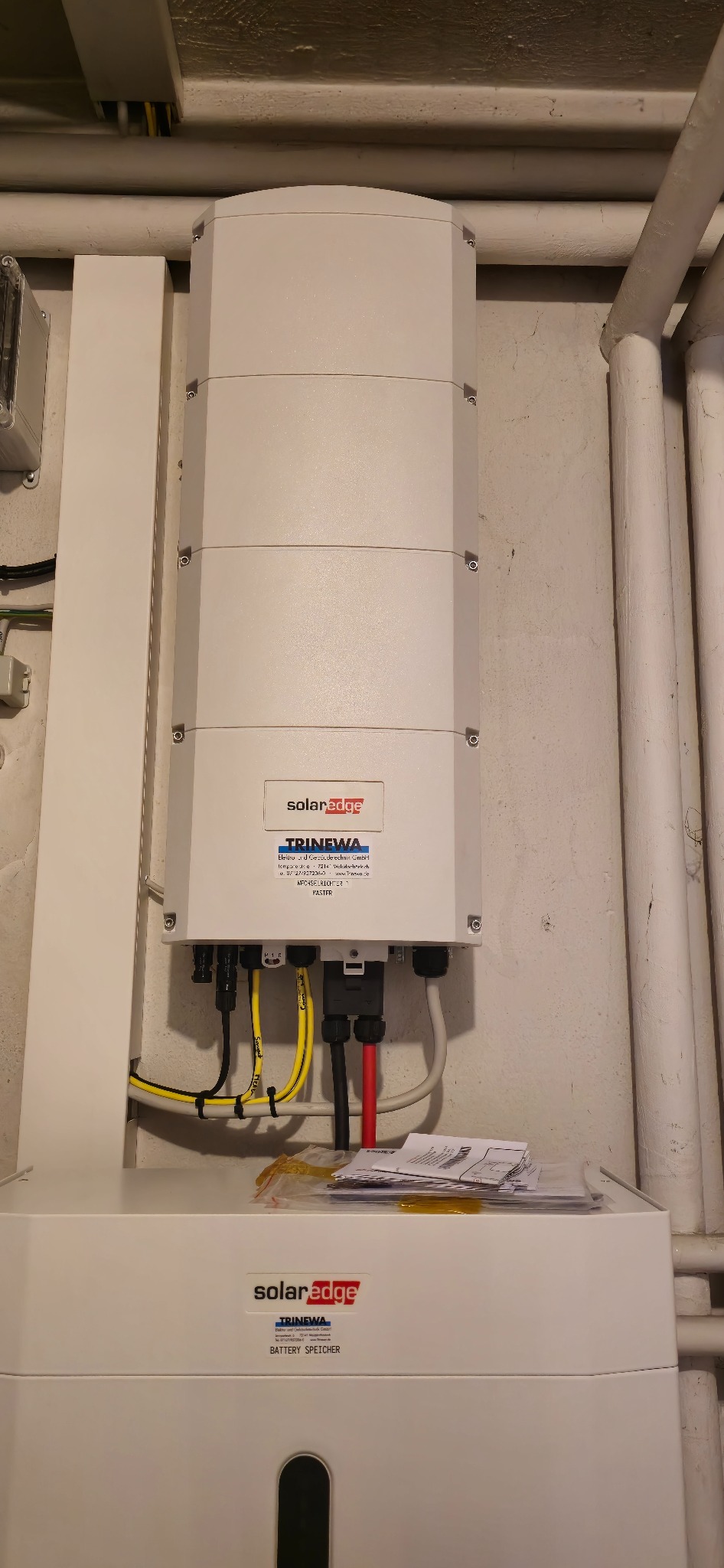 Zamontowany falownik SolarEdge Trinewa z podłączonymi kablami w Tuliszkowie. Widoczne rury instalacyjne na ścianie. Dokumentacja na górze urządzenia.
