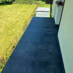 Wykończony taras z ciemnymi płytkami, widok z góry, z boku ściana budynku i zielony trawnik za metalową barierką.