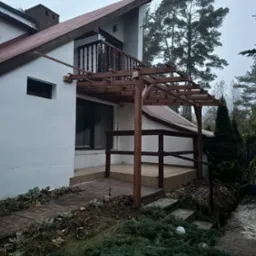 Drewniana konstrukcja zadaszenia tarasu w trakcie budowy, widoczne elementy szkieletu i podpory, dom z białymi ścianami i brązowym dachem w tle.