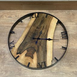 PIOTR PANFIL - Okrągły zegar ścienny z czarną obudową i tarczą z nieregularnego kawałka drewna o wyraźnych słojach, wskazówki czarne z niebieskim akcentem, położony na drewnianej podłodze.
