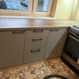 PIOTR PANFIL - Szare szafki kuchenne z czarnymi uchwytami pod drewnianym blatem, widok na okno z zielenią za nim, fragment odkurzacza przemysłowego na podłodze.