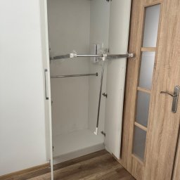 PIOTR PANFIL - Wnętrze białej szafy wnękowej z zamontowanymi drążkami na ubrania, częściowo jeszcze zabezpieczonymi folią, widoczne na tle drewnopodobnej podłogi i drzwi.