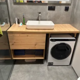 Nowoczesna łazienka z drewnianym blatem pod umywalką, pod którym znajduje się półka z ręcznikami i pralka automatyczna.