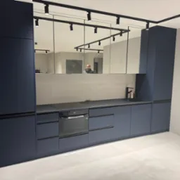 Nowoczesna kuchnia w odcieniach granatu z czarnym blatem i oświetleniem szynowym, odbicie osoby w lustrzanym panelu nad blatem.