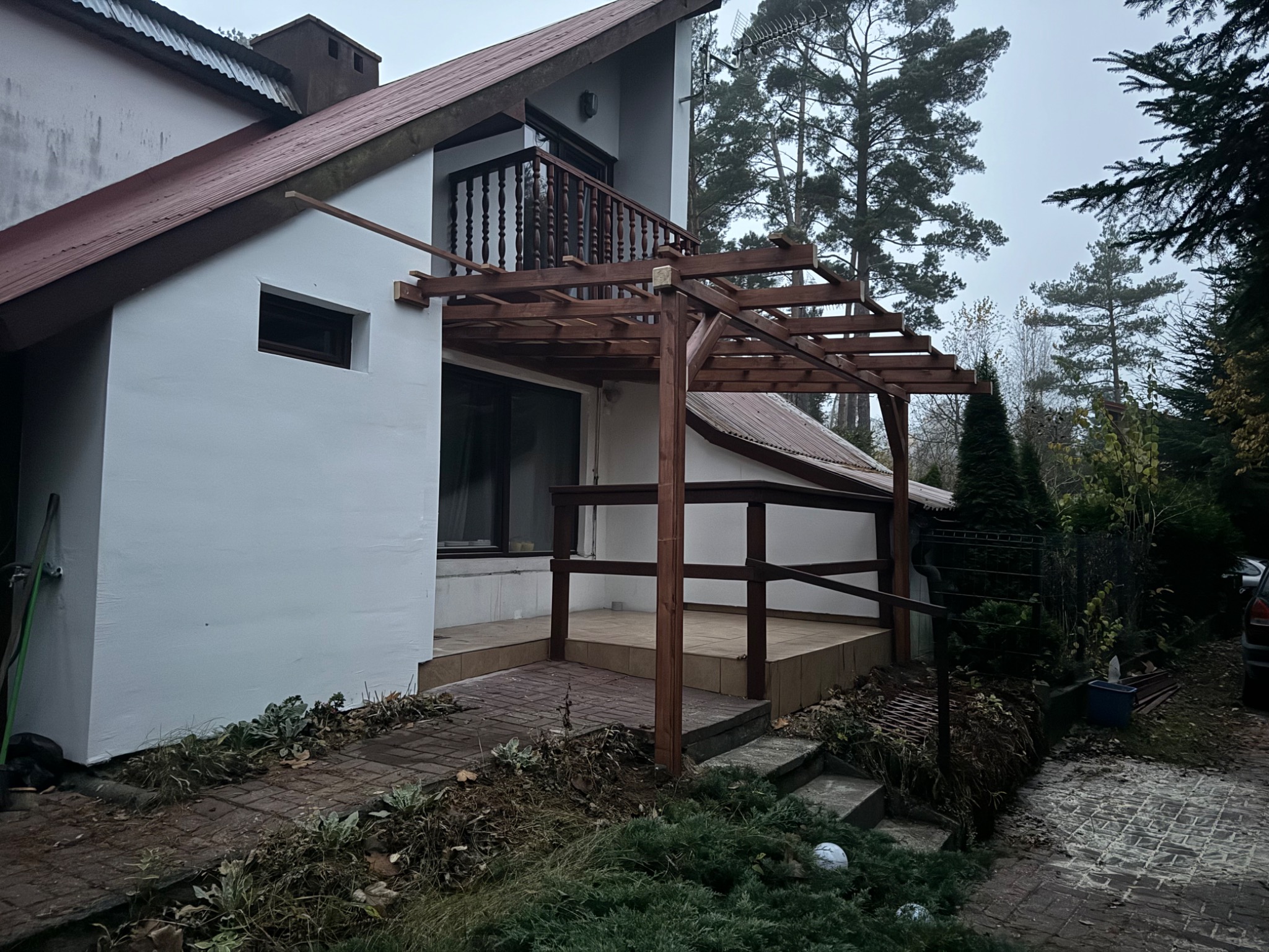 Drewniana konstrukcja zadaszenia tarasu w trakcie budowy, widoczne elementy szkieletu i podpory, dom z białymi ścianami i brązowym dachem w tle.