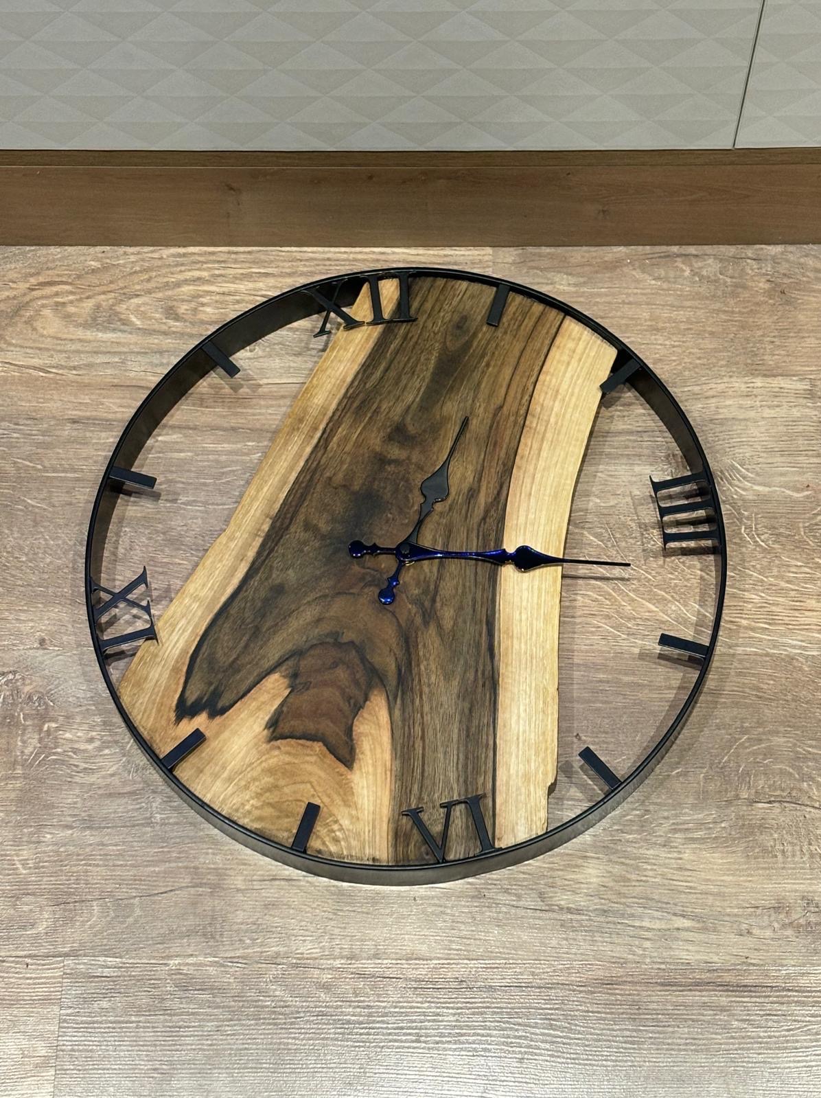 Okrągły zegar ścienny z czarną obudową i tarczą z nieregularnego kawałka drewna o wyraźnych słojach, wskazówki czarne z niebieskim akcentem, położony na drewnianej podłodze.