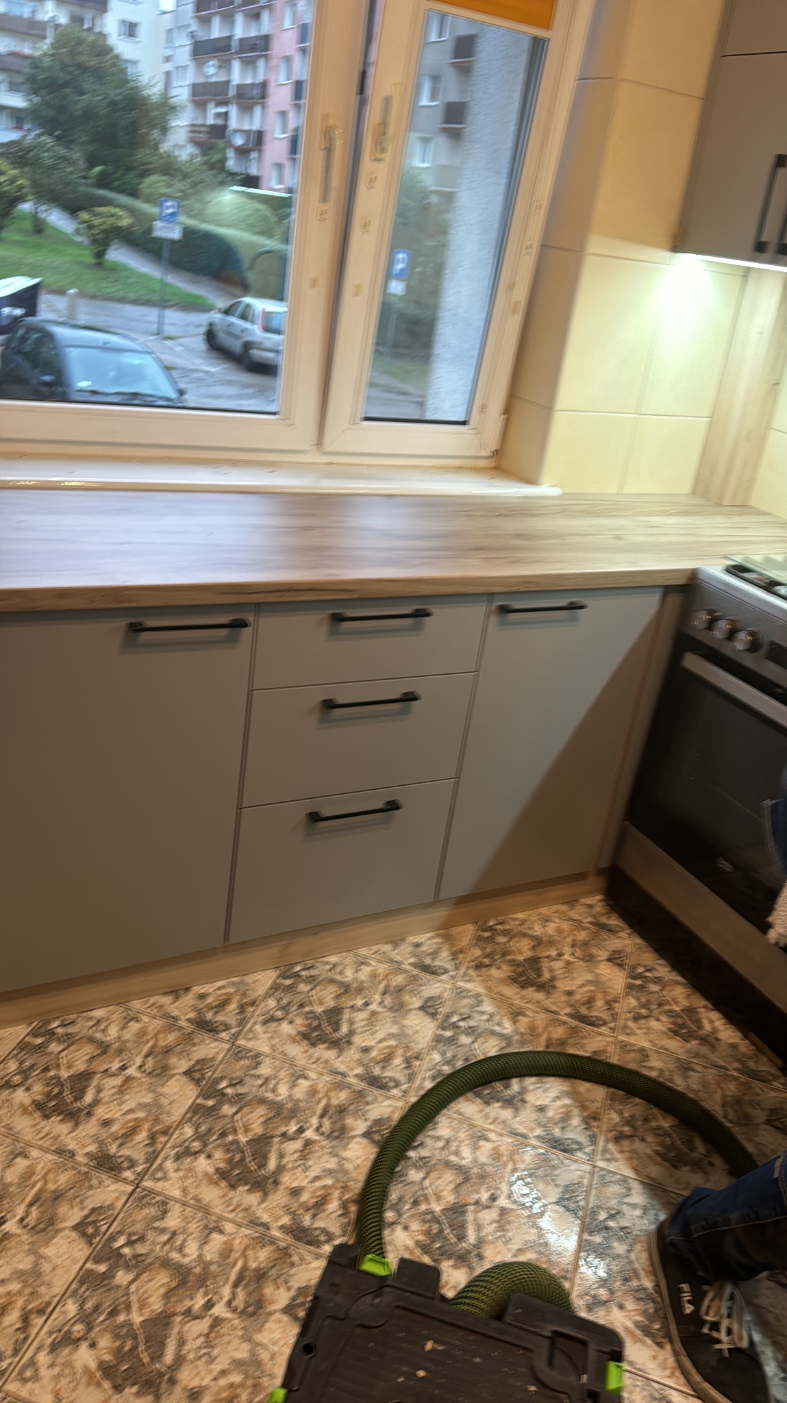 Szare szafki kuchenne z czarnymi uchwytami pod drewnianym blatem, widok na okno z zielenią za nim, fragment odkurzacza przemysłowego na podłodze.